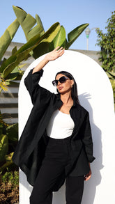 A woman posing with a black buttoned shirt, with the sleeves rolled up to the elbows, against a white circular backdrop with green foliage in the background.