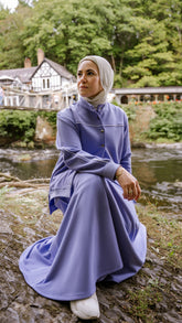 Woman in a purple dress sitting on a log by a stream with trees and a building in the background