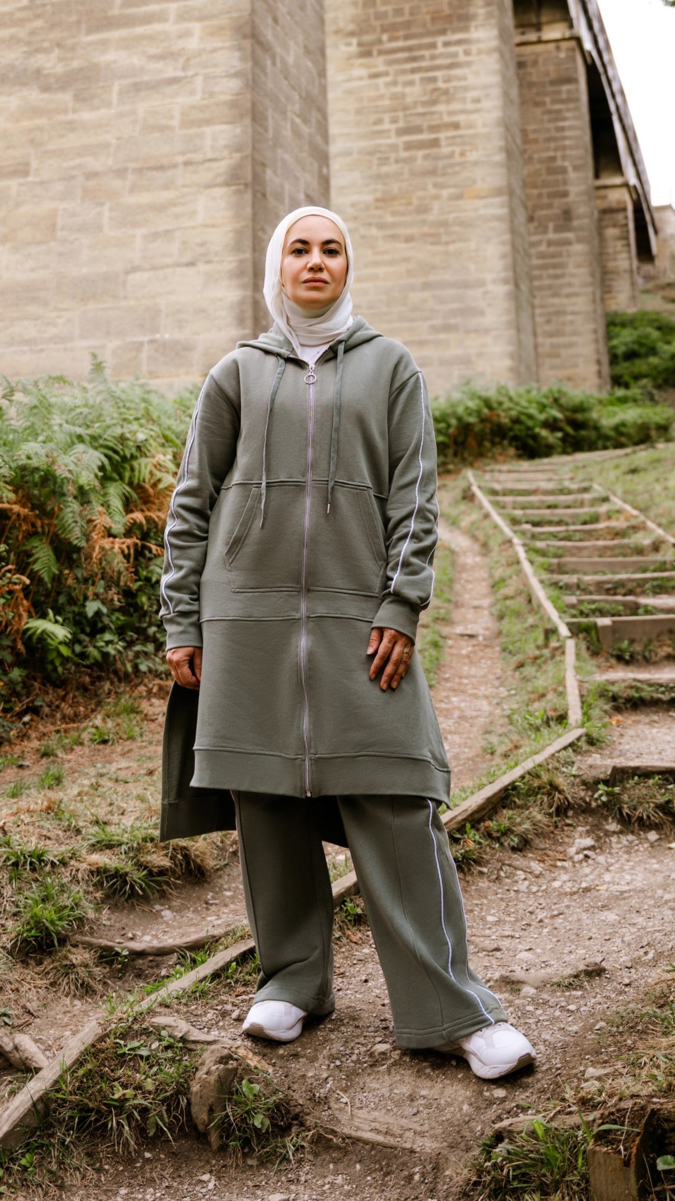 Person wearing a gray outfit standing on steps near a stone bridge with greenery.